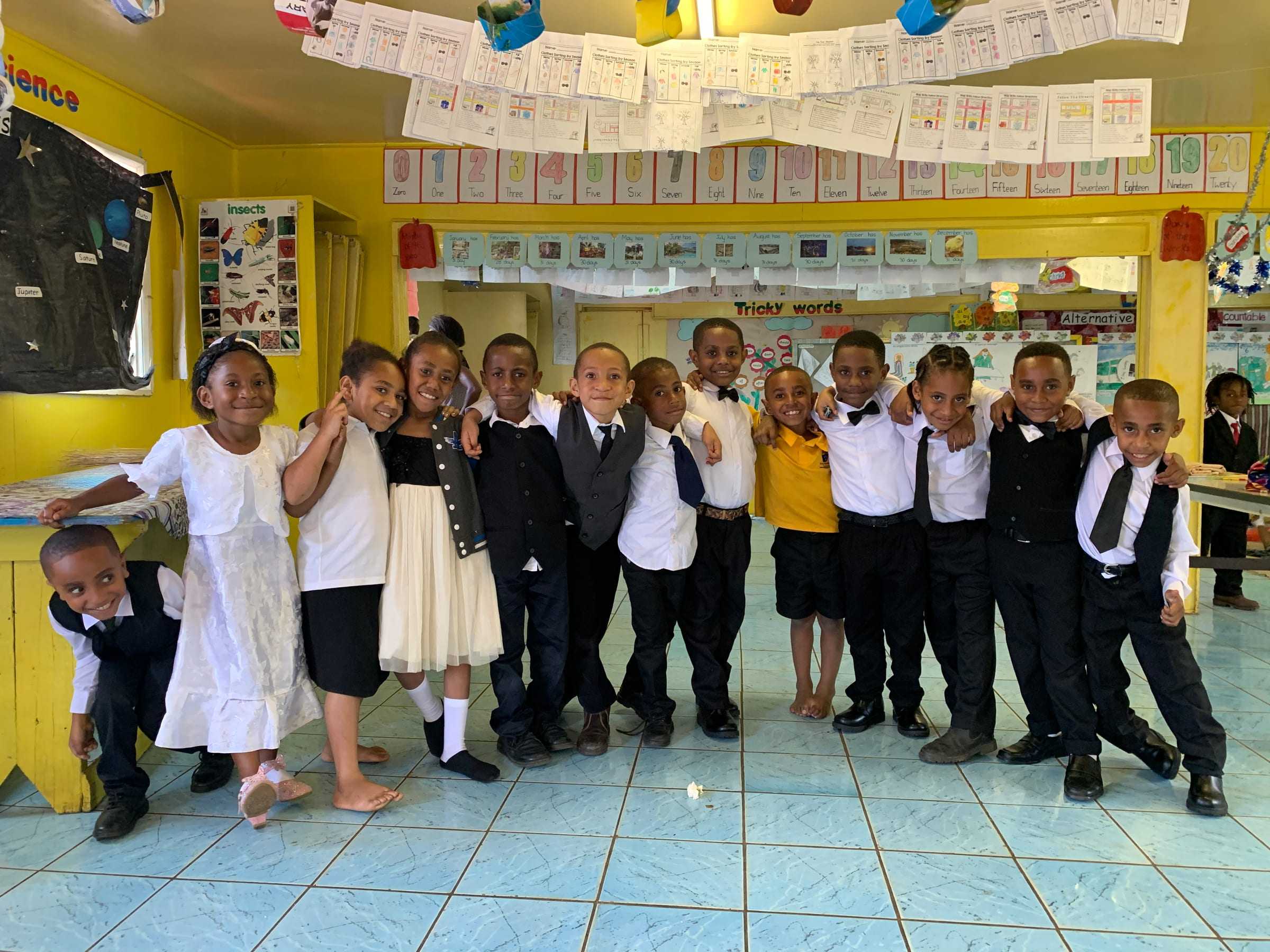 Early Learning children in white shirts and dresses smiling together in their bright yellow classroom.