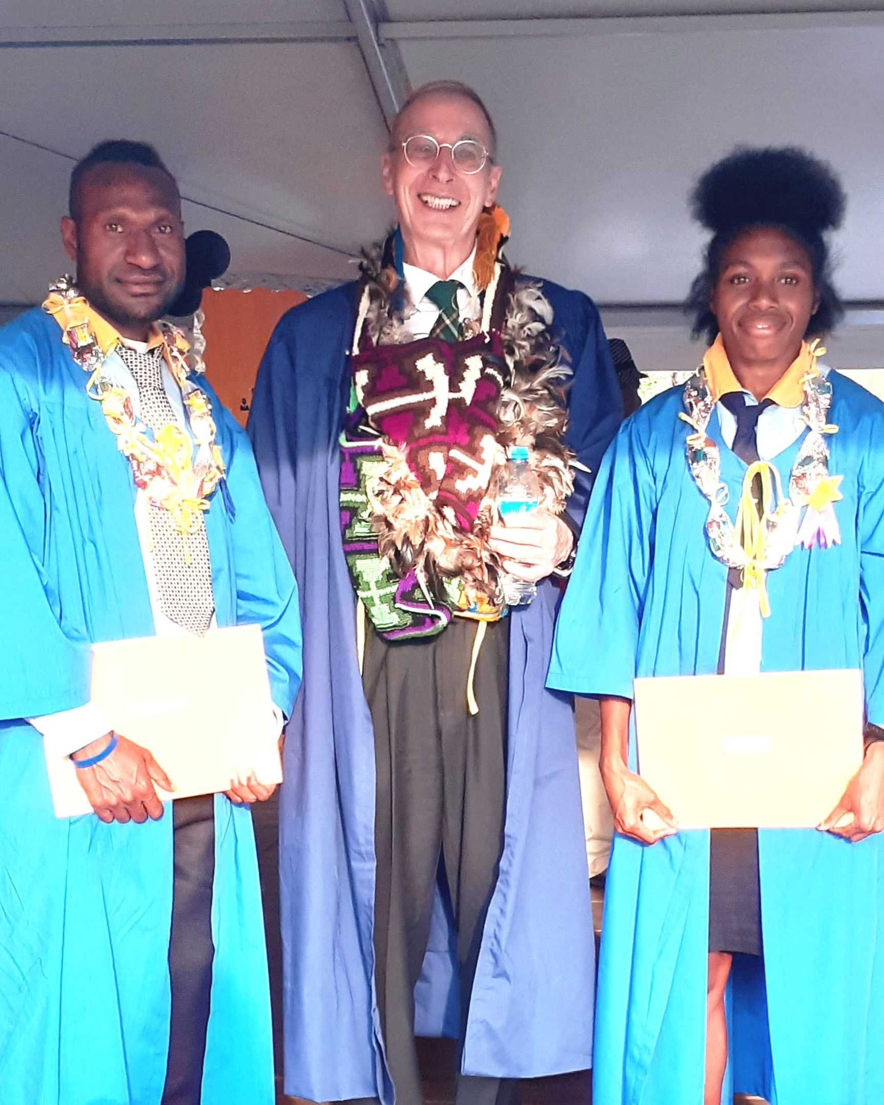 Two graduating students in blue gowns and garlands with the Director of Education.