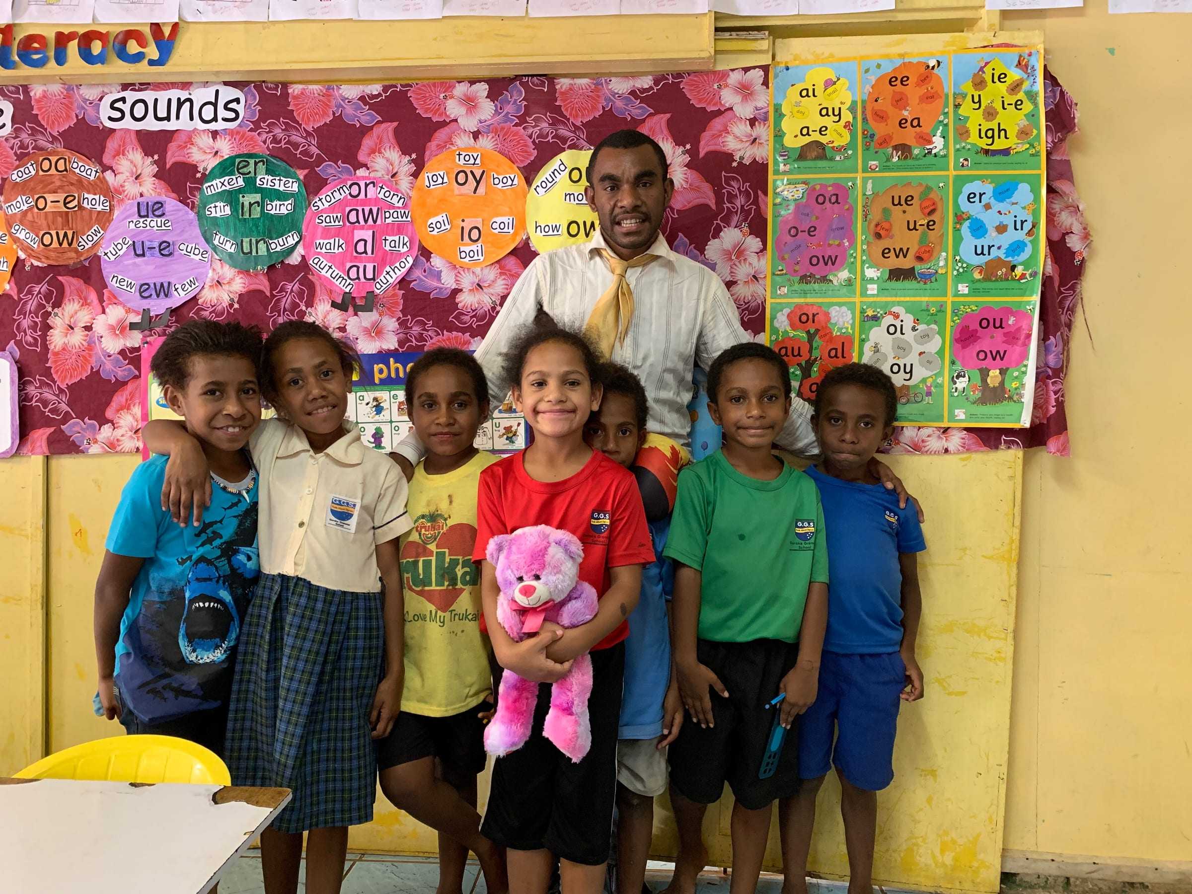 Primary children with their teacher in a classroom decorated with word charts and artwork.
