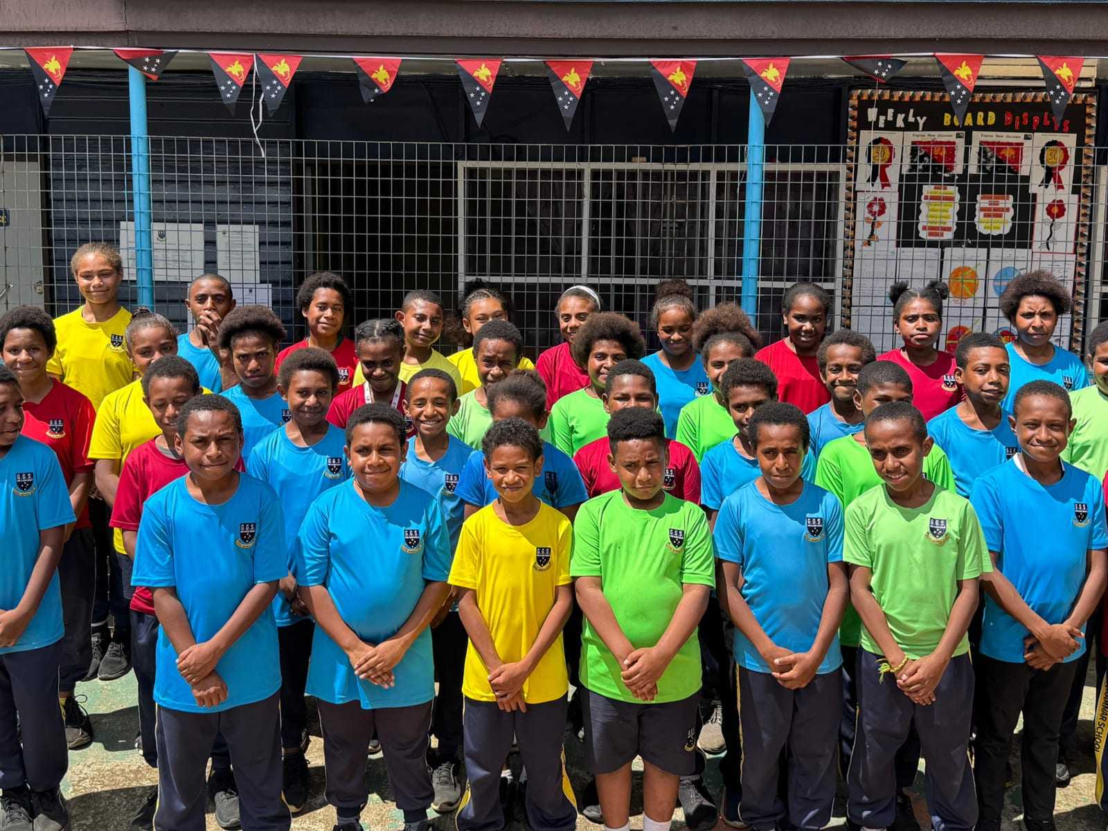 Primary students in bright house-colour shirts standing in rows outside their classrooms.