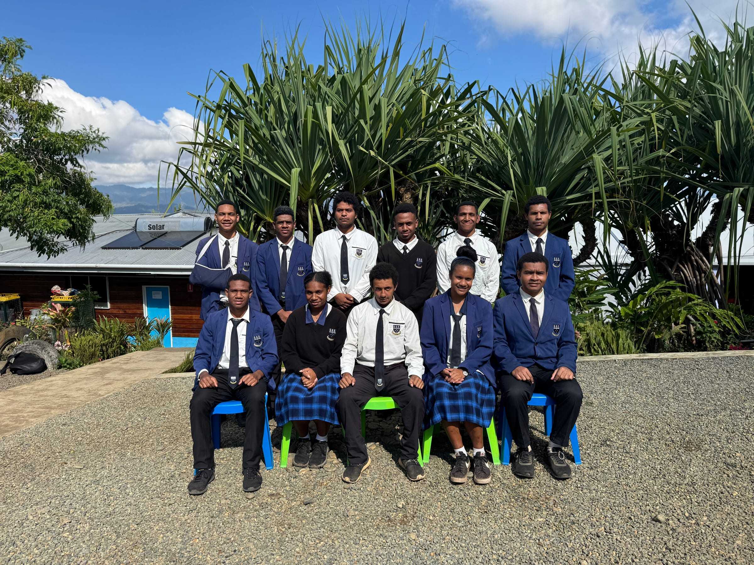 Senior students in GGS blazers and uniforms in the school garden under pandanus trees, with the Highlands behind.